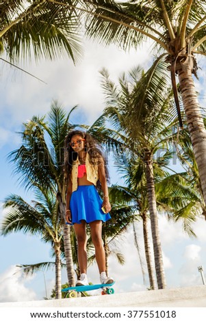 Outdoor lifestyle portrait of sporty black female in bright outfit and sunglasses. Young hipster girl ride her penny skateboard in front of palm trees. She is happy. Sunny summer day. Swag, fashion.
