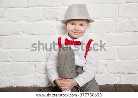 little boy wearing a red bow tie, suspenders and white shirtand against a white brick wall.