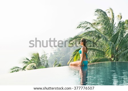 Young, beautiful female in cyan swimwear sitting in calm and peace poolside in the mountains.