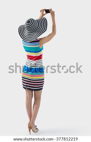 Vacation, tourism, travel concept. Mockup of unidentified woman wearing striped dress and hat taking a picture os selfie with her smartphone. Clipping paths for woman, smartphone and drop shadow