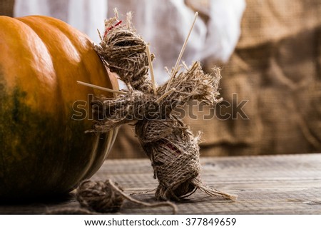 Closeup photo of terrible Halloween burlap voodoo puppet pierced with sticks on chest standing near orange pumpkin with clew of cord on table on white air fabric background, horizontal picture