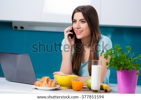 Picture a young woman eating breakfast and taking a phone call, sitting at the kitchen table.