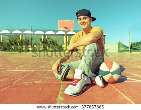 Young handsome guy sitting on basketball camp near the beach - Happy man take a rest after playing basket - Healthy lifestyle sport recreation concept - Vintage retro filter look 