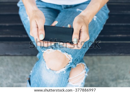 Girl in torn jeans sitting on a bench and uses a touch screen mobile phone. Top view close-up.