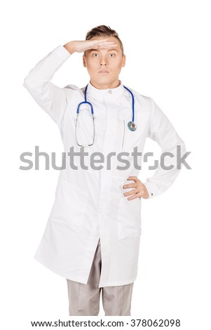 Portrait young male doctor in white coat looking away while keeping his hand over his eyes to protect himself from the sun rays. People and medicine concept. Image isolated on a white background.