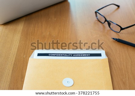 "Confidential Agreement" paper in brown envelope with black pen, eyeglasses and laptop on wood table background.