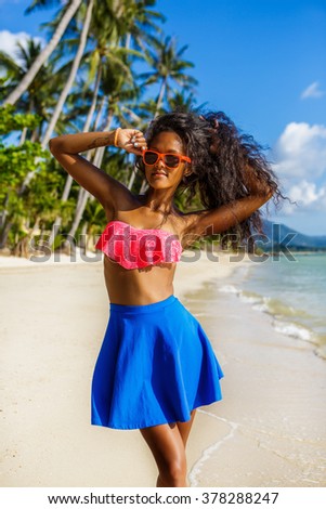 Outdoor lifestyle portrait of black teenage girl in pink bra and blue skirt. Young hipster woman correct her pink sunglasses and her curly hair. Sunny hot summer day at tropical beach with palms.