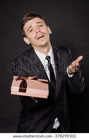Portrait young businessman in black suit looking at camera with gift box. Christmas, x-mas, winter, valentine's day, birthday, happiness concept. Image on a black background.