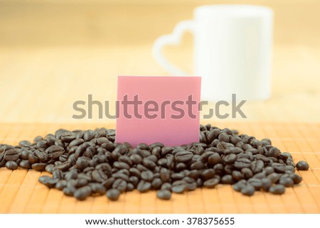 pink square paper note in raw coffee bean over wood floor with white mug as background