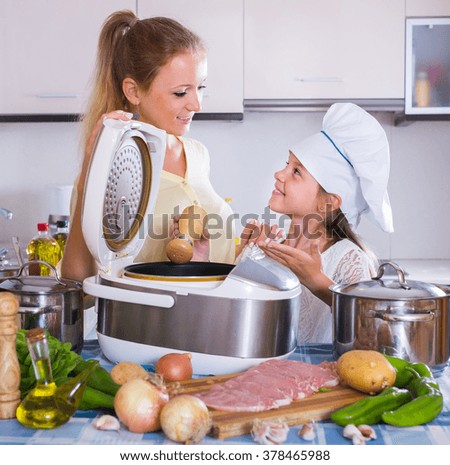 Smiling girl helping mom cooking vegetables and meat in multicooker at kitchen
