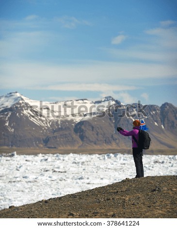 Woman traveller taking a photo of Jokulsarlon glacier Lagoon with iceland flag flapping in the wind, snowcapped mountains in the background