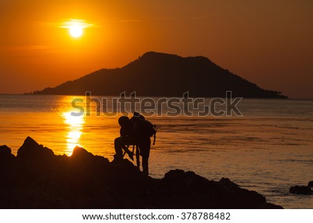 Silhouette of a male photographer standing on the rock at the beach of Ao Kra Ting Bay and preparing his equipment for sunset shot behind the island in the sea. (Koh Nom Sao)