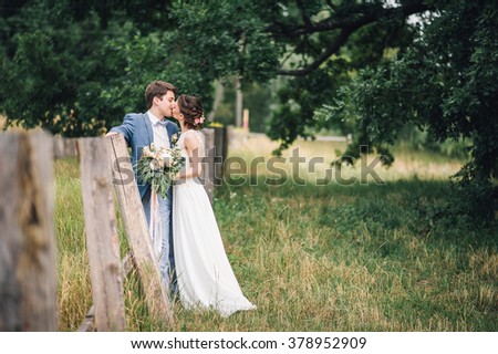 couple in wedding attire with a bouquet of peonies and greenery standing in the field near wooden fence with trees on background, the bride and groom 