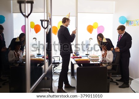Business woman celebrating birthday and doing a party with colleagues in her office. A friend with mobile phone takes pictures of her blowing out clandles on birthday cake. Wide shot