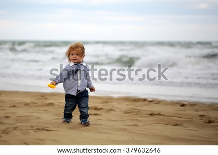 Cute fair-haired kid tiny little child baby boy in jeans striped shirt and tie running about and playing with sand on beach on windy murky day on blurred seascape background, horizontal picture 