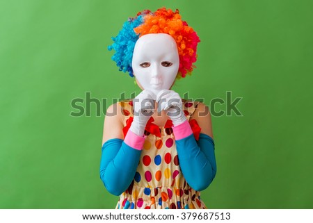 Portrait of a funny playful female clown in colorful wig holding a mask on her face, standing on a green background