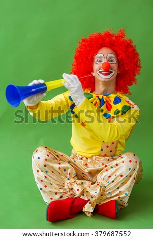 Funny playful clown in red wig holding a blowing horn, looking at camera and smiling, sitting on a green background
