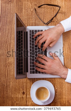 Top view of man's hands using a laptop and typing on a wooden background, a cup of coffee and eyeglasses on the side