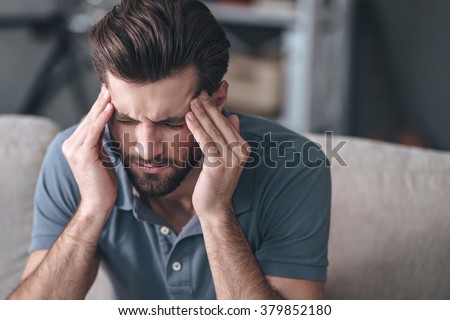 Feeling stressed. Frustrated handsome young man touching his head and keeping eyes closed while sitting on the couch at home Royalty-Free Stock Photo #379852180