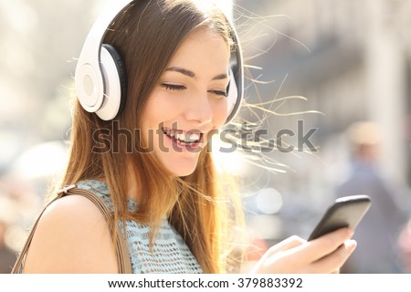Portrait of a happy girl listening music on line with wireless headphones from a smartphone in the street in a summer sunny day Royalty-Free Stock Photo #379883392