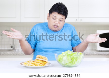 Picture of fat person sitting in the kitchen and looks confused to choose hamburger or salad