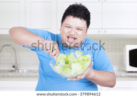 Picture of cheerful overweight man sitting in the kitchen while eating a bowl of fresh vegetable salad