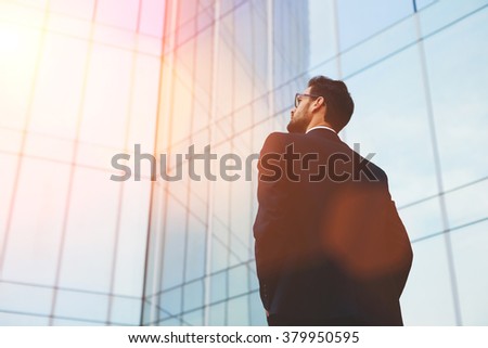 Back view of serious businessman in eye glasses looking on copy space while standing against glass skyscraper, young professional employee waiting for international partners outdoors near big company Royalty-Free Stock Photo #379950595