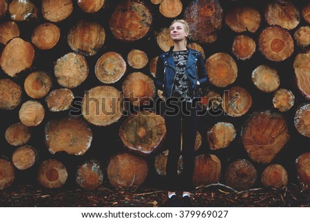 Trendy young woman admires something while standing outdoors against stacked wooden logs background, pretty hipster girl dreaming about good life while waiting for someone outside near cut tree trunks