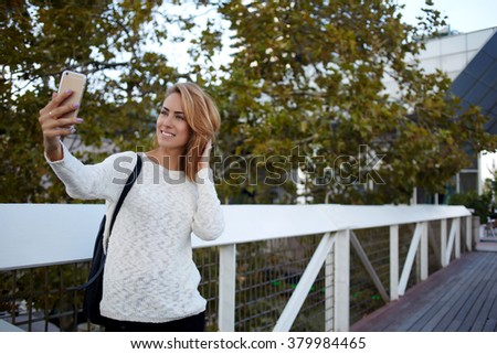 Happy female tourist photographing herself via cell telephone camera while standing on a bridge near house, cheerful European woman shoots video on mobile phone while strolling in spring day outdoors