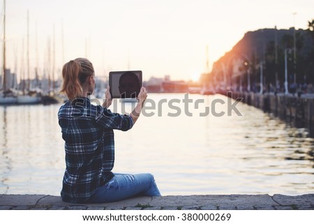 Back view of a young female making photo on digital tablet while sitting against beautiful landscape background with copy space, woman photographing view with touch pad camera while relaxing near sea 