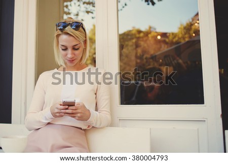 Gorgeous young woman searching information on mobile phone while sitting in modern coffee shop, attractive female student using her cell telephone while relaxing after lectures in University