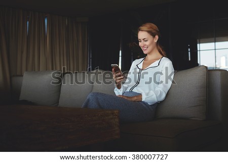 Young female with beautiful smile chatting on mobile phone while sitting in comfortable modern restaurant, businesswoman reading good news on cell phone from her partner while enjoying rest in cafe