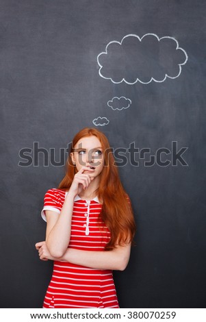 Thoughtful pretty young woman standing and thinking over  blackboard background with blank bubble 