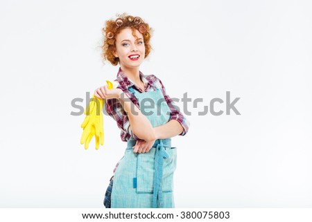 Cheerful beautiful young housewife with red curly hair in blue apron holding yellow rubber gloves over white background