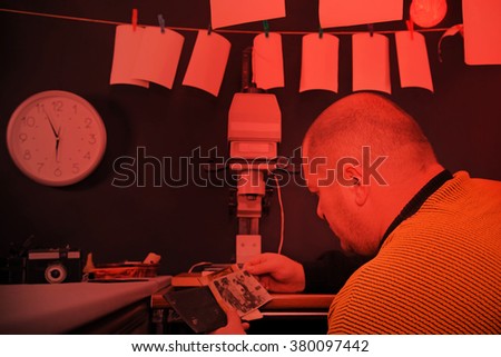 Retro darkroom in red light. Adult photographer looking at old black and white photo.