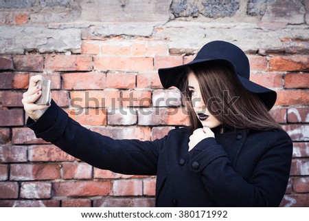 Stylish teenage girl with black lipstick and black hat taking self portrait next to an old brick wall. Toned effect