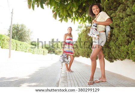 Two diverse joyful teenagers girls friends together in suburban home exterior street with roller blades in sunny outdoors. Sporty action living. Adolescents enjoying holiday, smiling looking.