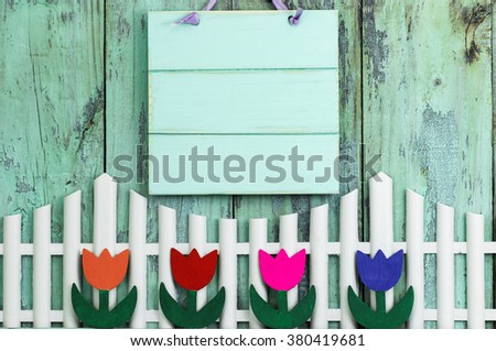 Blank mint green wood sign hanging over white picket fence with row of colorful spring flowers on antique rustic wooden background; purple, pink, orange, red tulips