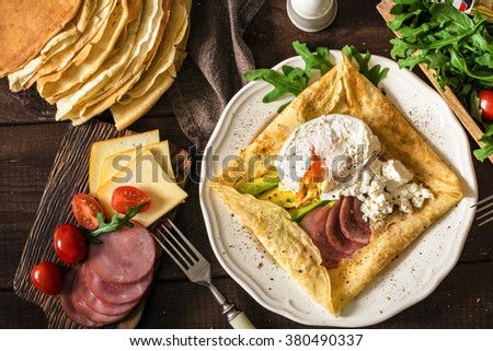 Crepe galette with ham, avocado, soft white cheese and poached egg on white plate. Sliced yellow cheese, pastrami, cherry tomatoes, green salad and stack of crepes on side. Top view