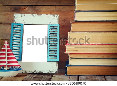 stack of old books next to decorative sailing boat and blank frame wooden table. vintage filtered image. template, ready to put photography
