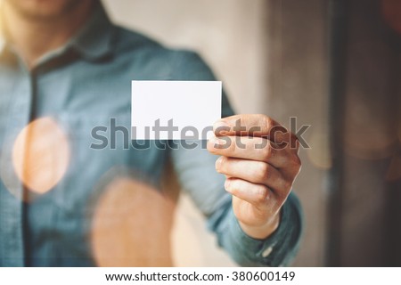 Man wearing blue jeans shirt and showing blank white business card. Blurred background. Horizontal mockup