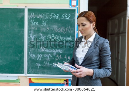  inside a school class. Beautiful red-hair teacher is standing  in-front a school board. She is holding a copybooks. Teacher is wearing gray jacket and white blouse