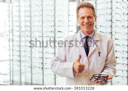 Handsome middle aged ophthalmologist offering eyeglasses, showing OK sign and smiling while standing in his office