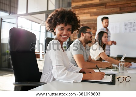 Portrait of happy young african woman sitting at a business presentation with colleagues in boardroom. Female designer with coworkers in conference room.