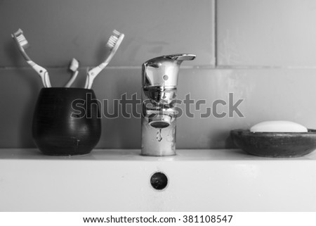 Scarce water/Black and white image of a bathroom sink with the tap dripping a drop of water, two normal toothbrushes plus a small one and a piece of soap in a soap dish.