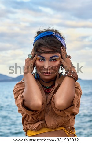 Asian Woman with bright fashion makeup, ethnic clothes in boho style, seating near the sea. Beauty photo shoot in ethnic fashion style.
