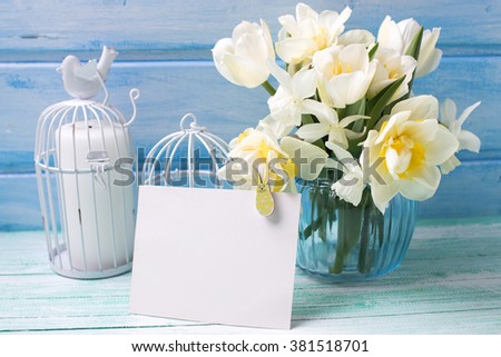 Bright white daffodils and tulips  flowers in blue vase, candles  and empty tag  on turquoise  painted wooden planks against blue wall. Selective focus.