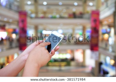 hand hold and touch screen smart phone, on blurred photo of  department store shopping mall center and people background