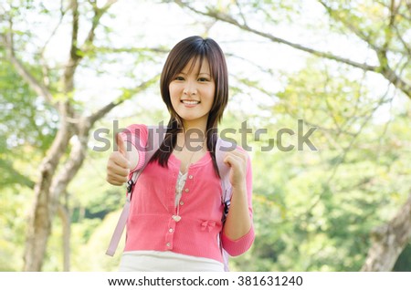 Young Asian university girl student standing on campus lawn, showing thumb up and smiling.