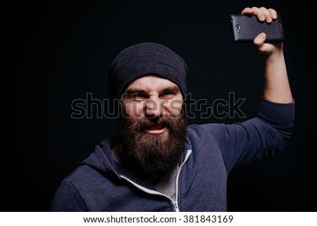 Beautiful young man with a beard in a gray hoodie makes self, smiling, studio shot on black background, aggressive guy throws the phone
